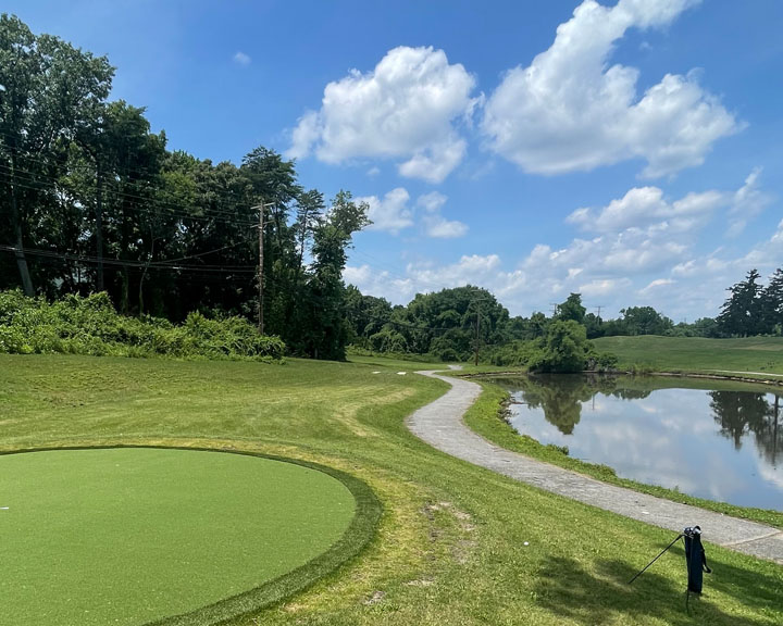 A narrow paved path follows the edge of a body of water. On the other side of the path is a circular putting green.