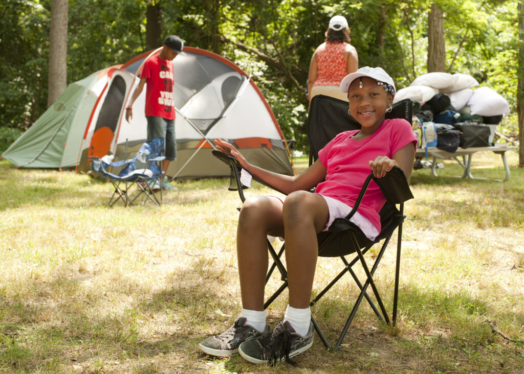 A smiling child sits in a folding camp chair in the grass. Behind her are several dome tents and a picnic table piled with bags and pillows.