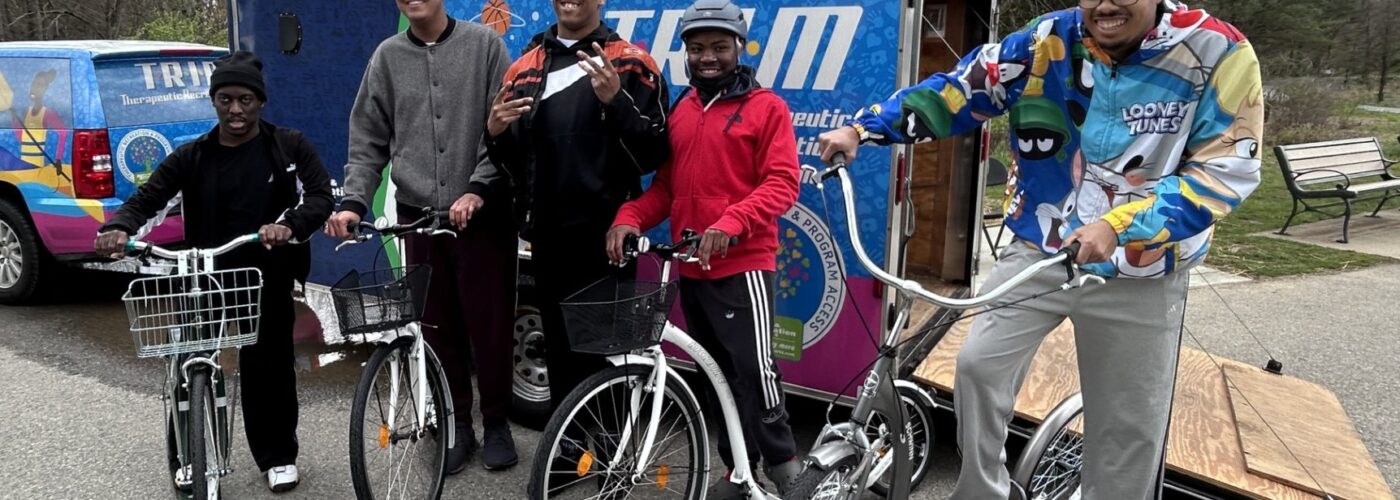Four young men pose with kickbikes in a parking lot, near a trailer branded with a colorful logo and images.