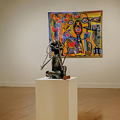 A gallery with an abstract sculpture on a white podium. Behind it on the wall is a colorful abstract painting.