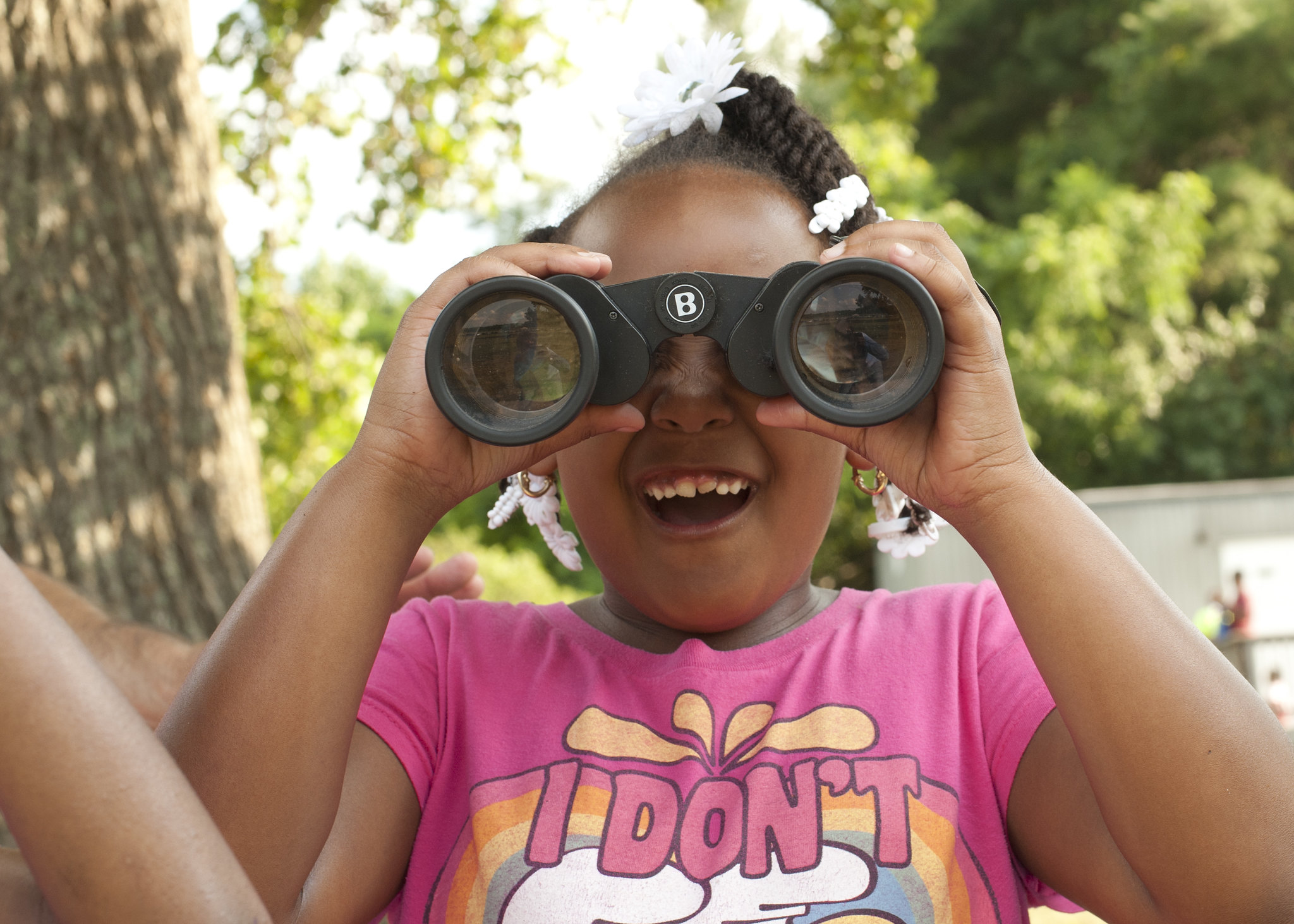 A child in a pink t-shirt uses black binoculars to look directly at the photographer of the image. Her mouth is open in an expression of delight. Behind her is a blurry tree.