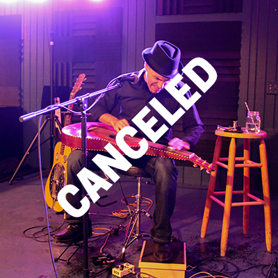A seated man plays a guitar-like instrument that is lying flat across his lap. Across the image is word CANCELED.