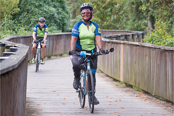 A woman rides a bike along a curved, wooden bridge. She's wearing a helmet and a biking shirt. There is a man behind her wearing the same shirt also riding a bike.