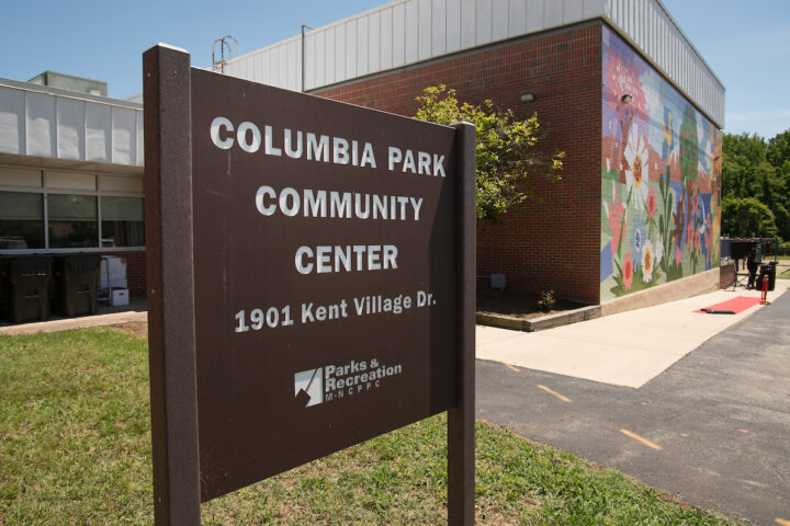A brown sign reading COLUMBIA PARK COMMUNITY CENTER is mounted in the grass in front of a large, brick and glass building. One wall of the building is covered by a colorful mural.