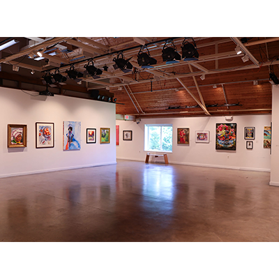 A room with white walls, solid floors, and a vaulted wooden ceiling. There is a window against the back wall casting a reflection on the shiny floor. There are various works of art hanging on the walls.
