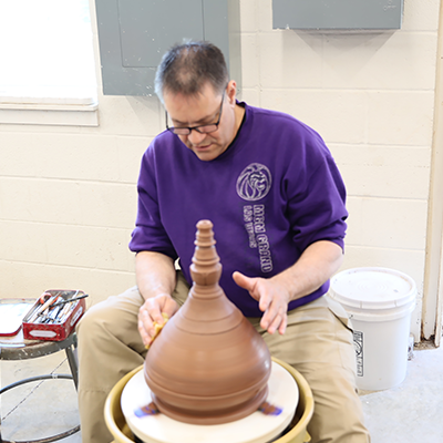 A man in glasses and a purple shirt shapes clay into a large conical vessel on a spinning pottery wheel.