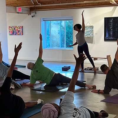 Inside an art gallery, adults lying on yoga mats execute a stretch, led by an instructor standing at the front of the room.