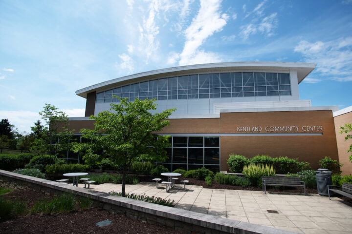Exterior view of a large brick, glass, and metal building with a landscaped patio. Letters on the building read KENDLAND COMMUNITY CENTER. There are benches on the patio as well as cafe-style tables with stools.