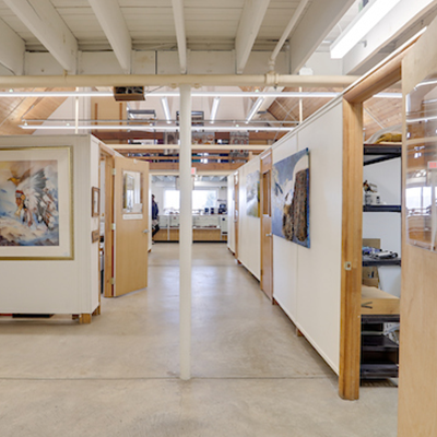 View down a hallway with a cement floor and wooden doors to multiple artists' studios along both walls. Framed artworks hang on the walls.