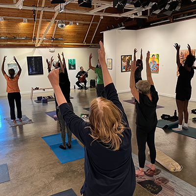 Inside an art gallery, adults stand on yoga mats being led in a stretch by an instructor. Next to the instructor is a seated man playing the guitar.