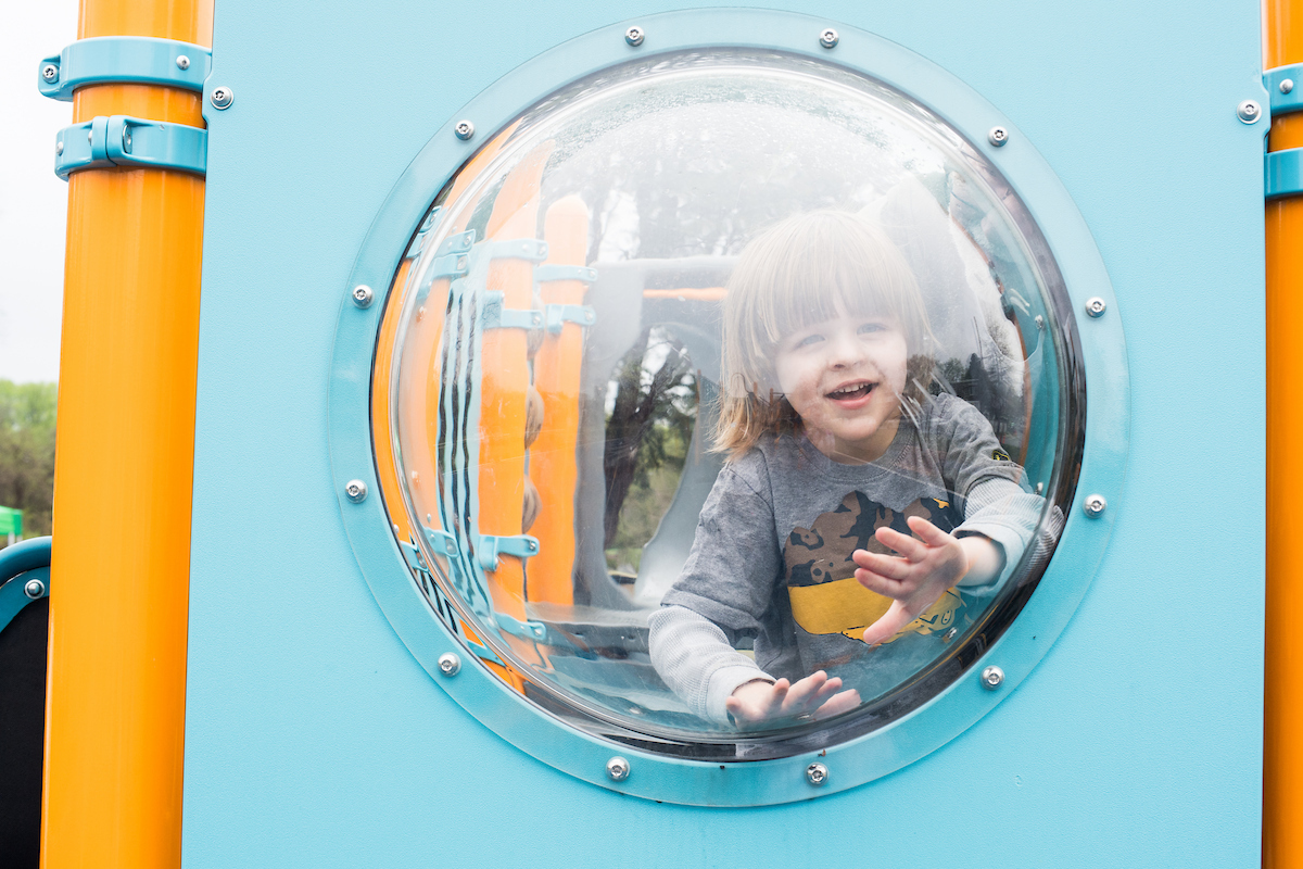 A small child smiles from behind the plexiglass bubble-window feature of an outdoor playground. The play equipment is sky blue with yellow vertical posts.