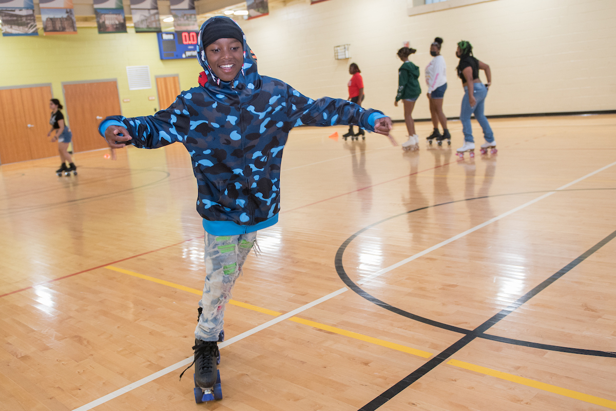 A smiling youth in a blue hoodie roller skates in a gymnasium. Behind him other teens are roller skating.