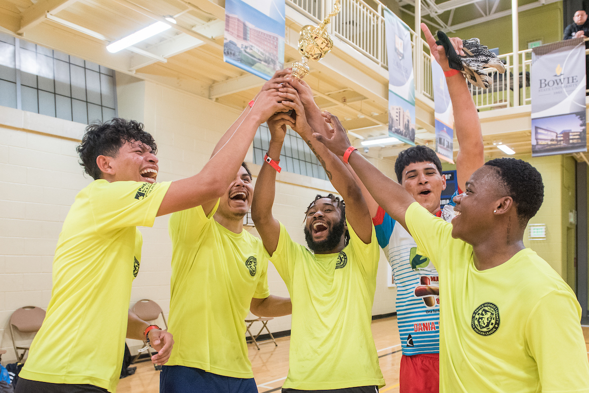 In a gymnasium, five young men celebrate while holding a golden trophy high above their heads. Their mouths are open and their faces show extreme joy and emotion. A spectator watches from an elevated walking track around the perimeter of the gym.