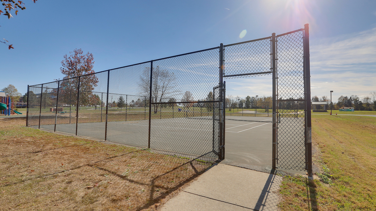 A sunny, outdoor tennis court surrounded by a black chain-link fence. Beyond the court is a large grassy field. To the left, an outdoor playground is just visible.