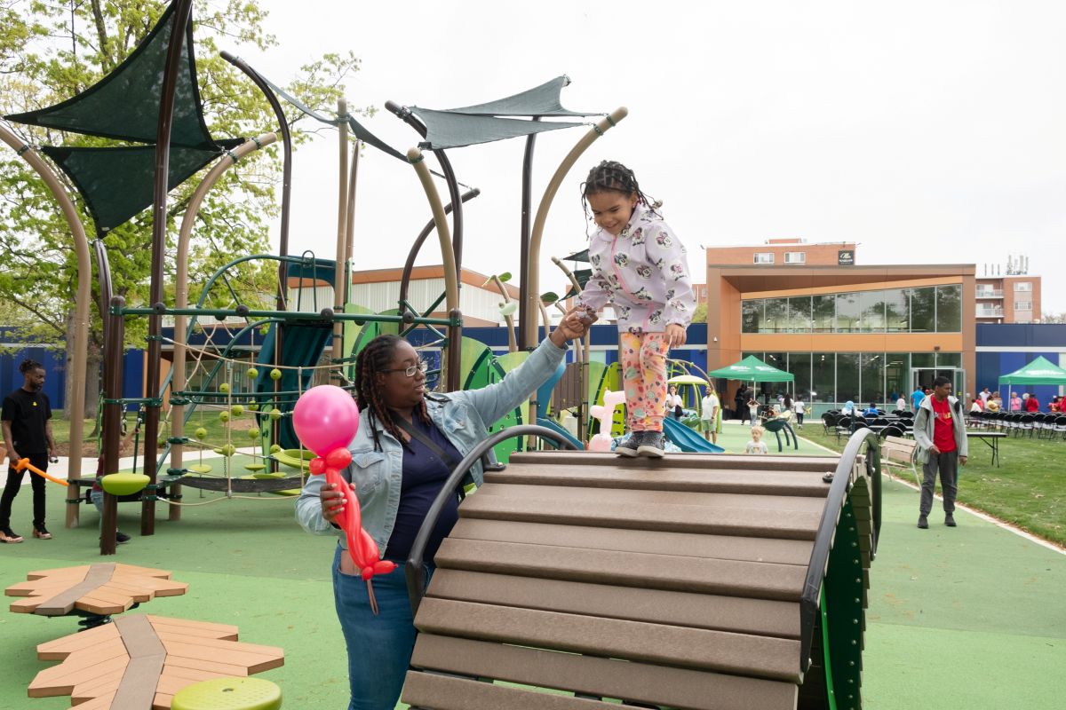 A mom holds her daughter's hand as the daughter climbs on a tall piece of playground equipment shaped like an arch. Behind them is a large outdoor playground that is mostly green. In the background is a large, modern, brick, glass, and metal building with sections that are royal blue.