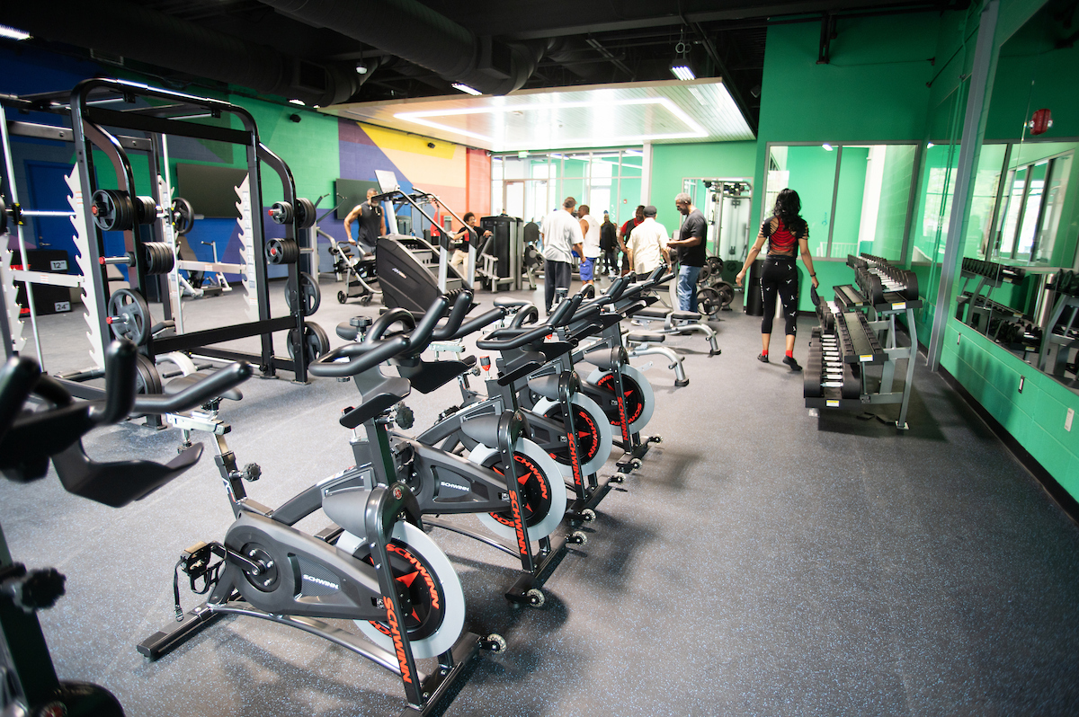 A large fitness room containing free weights, stationary bicycles, and a number of pieces of exercise equipment. The room has green walls, with large geometric shapes in purple, orange, and yellow, painted on the far wall.