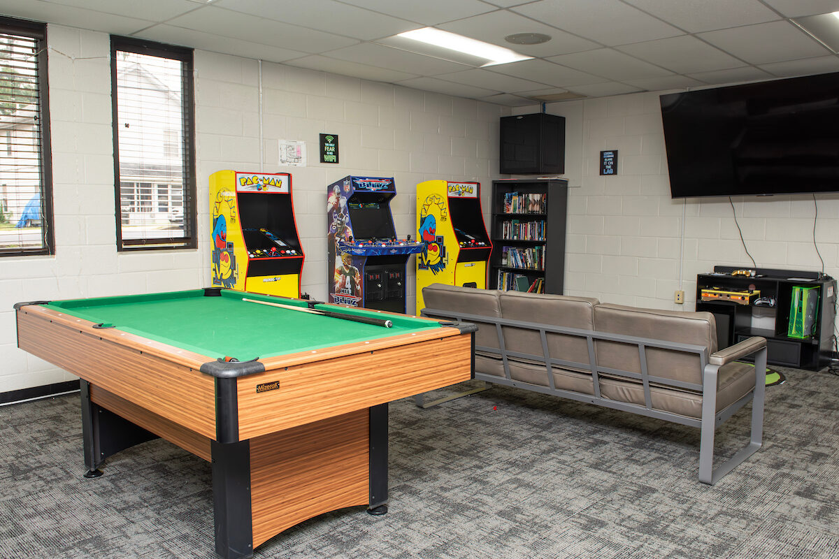 A carpeted game room containing a pool table, a gray sofa, a large tv with multiple gaming consoles below it, a bookcase filled with DVDs, and three arcade games, including Pac-Man.