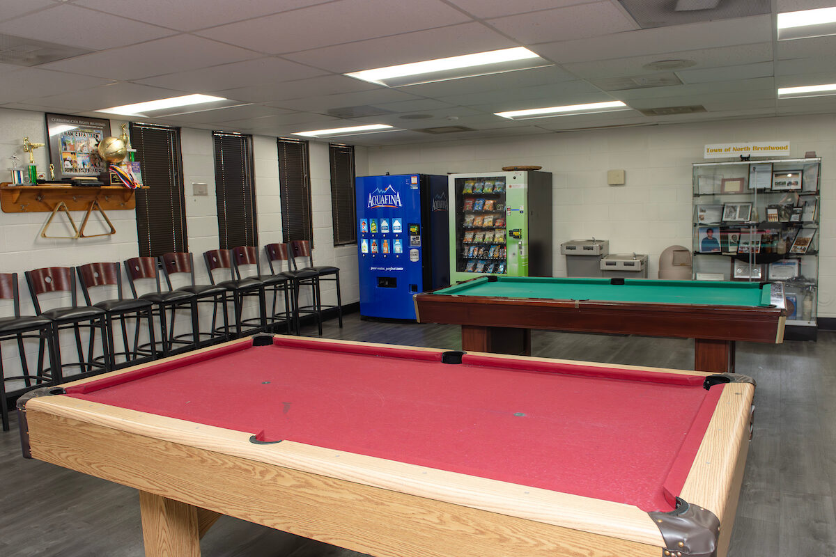 In a lounge area are two pool tables, one covered in red felt and one covered in green felt. Along the left wall is a row of stools, and a shelf containing several trophies. Along the rear wall are two vending machines, and a glass case full of framed photos and certificates, along with two water fountains.