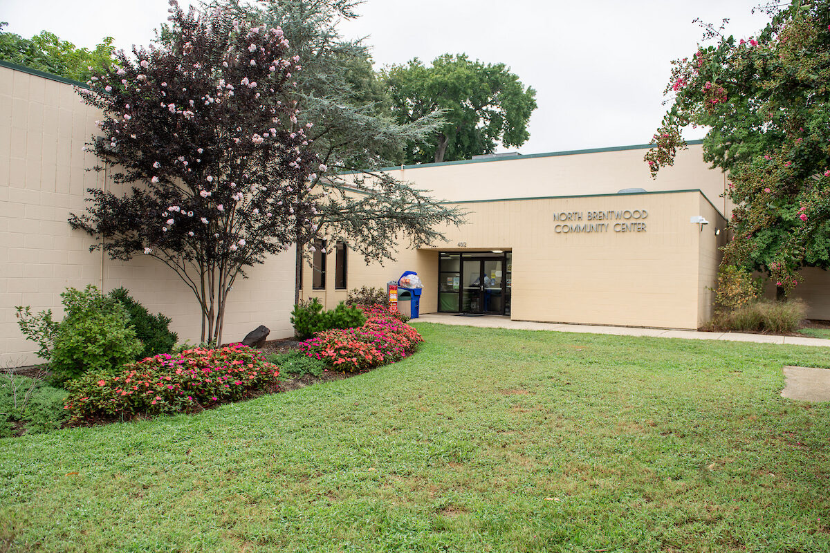Exterior view of the entrance to a large, beige, cinderblock building with green trim. Letters on the building spell out NORTH BRENTWOOD COMMUNITY CENTER. There is a sidewalk leading to the entrance doors, next to a lush, green yard with landscaped flowerbeds and several flowering crepe myrtle trees.