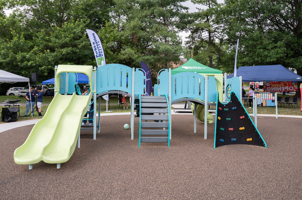 A play structure at an outdoor playground, featuring slides, a climbing wall, bridges, platforms, monkey bars, and more. Beyond the edge of the playground, event tents are set up in the grass under some tall trees.