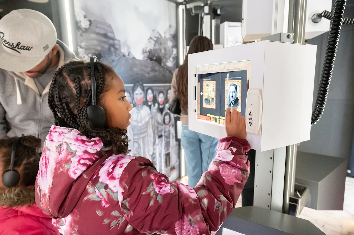 A girl in a floral coat interacts with a video monitor museum exhibit, while wearing headphones connected to the exhibit. Behind her other visitors explore the museum.