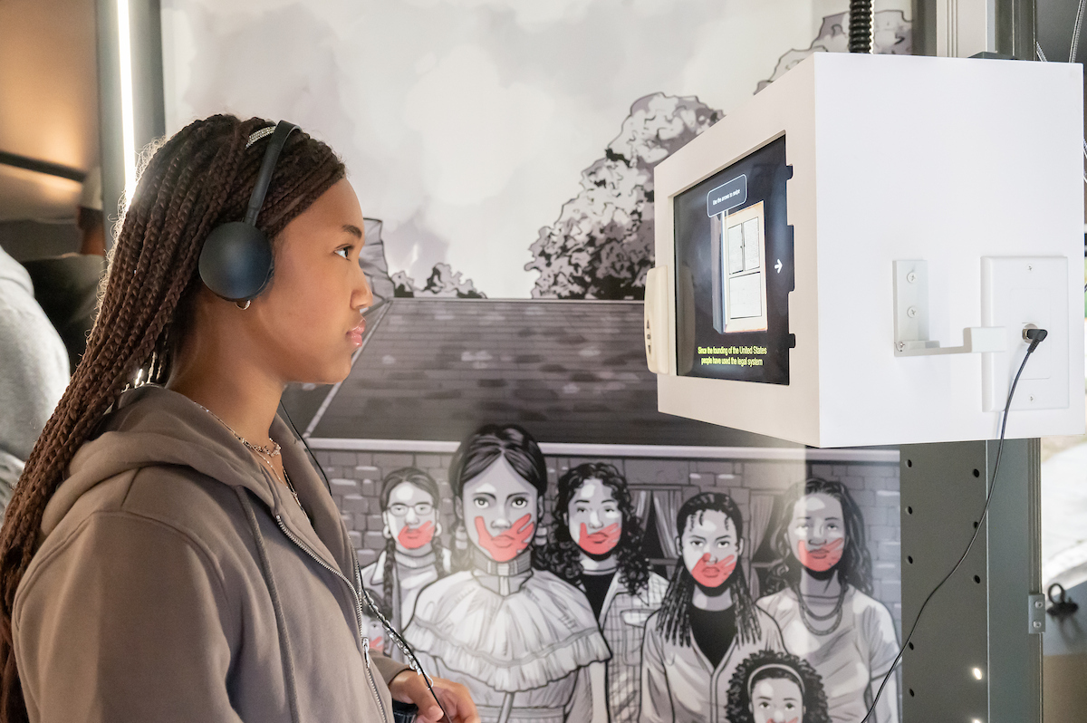 A woman with long braids looks at the interactive monitor of a museum exhibit, while wearing headphones connected to the exhibit. Behind her is an illustration of women from various historic eras with red hand marks painted across their mouths.