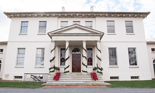 Exterior view of a large, white, two-story, Colonial-style mansion with four columns supporting the front porch. The columns are wrapped with evergreen garland, and there are red wreaths on the two windows flanking the front door. Potted poinsettias line the steps to the entrance.