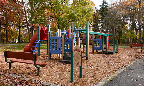 A colorful playground with multiple climbing structures, platforms, slides, and ladders. Several red benches are installed near the play equipment, and the ground is littered with fallen leaves. In the background is a line of tall trees that are beginning to change color for fall.