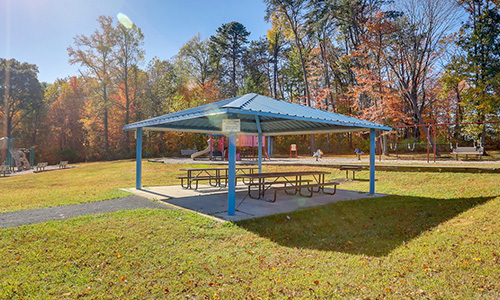 A square picnic shelter with a metal roof covering two long picnic tables that sit on a concrete patio in the grass. Behind in the distance is a playground, and a line of tall trees whose leaves are beginning to change color to red and orange.