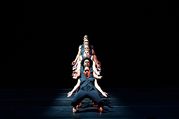 Seven barefoot dancers in matching black pants and black sleeveless shirts stand in a single file line on a dark stage, with their arms out to their sides, palms upward. All their faces are visible. The dancers in front bend their knees so they are low to the ground in a limbo-like pose, while the dancers at the back of the line are standing.