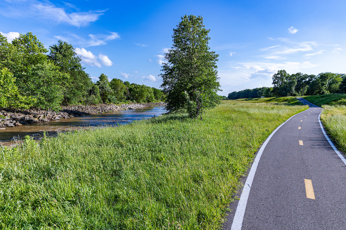 A paved asphalt trail with a dotted line down the middle cuts through a grassy field and curves up a slope toward some trees. On the left is a brown river or creek.