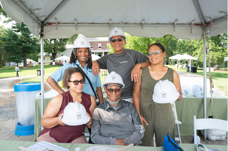 Five people wearing white hard hats pose together beneath an event tent at a community gathering.