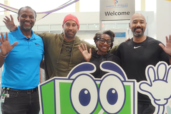 Four adults stand and wave behind a cut-out image of a green and white square mascot with eyes and a waving arm. The man on the left is Director Darius A. Stanton.
