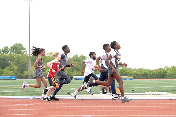 Several kids running around a track. They are all wearing athletic shirts and shorts. It is a bright cloudy day.