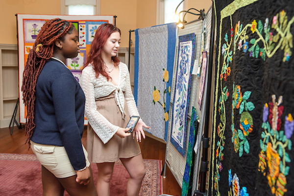 Two women looking at challenge quilts that are hanging on the wall. They picture various flowers, and designs.