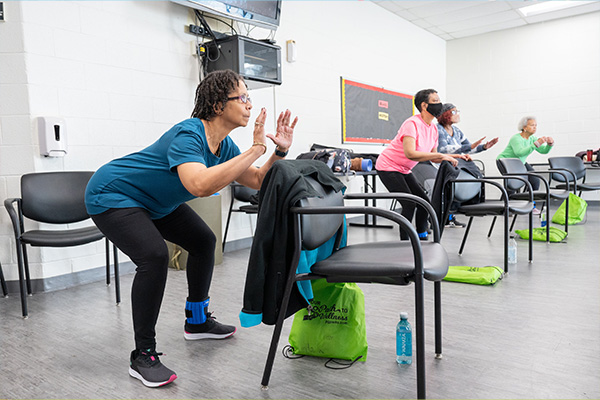 Seniors in exercise clothes crouch behind black chairs with their arms slightly raised.