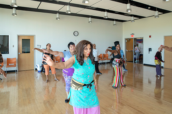 About ten adults dance in a large, multipurpose room, wearing special belts around their hips adorned with silver jingles.