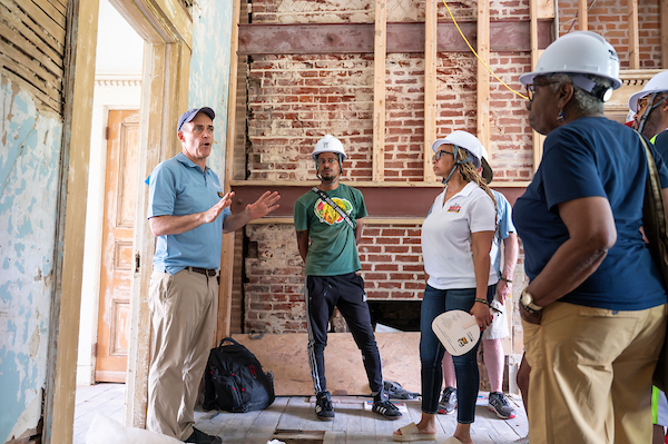 Five people wearing hard hats listen to a man speaking in a house that is under construction.