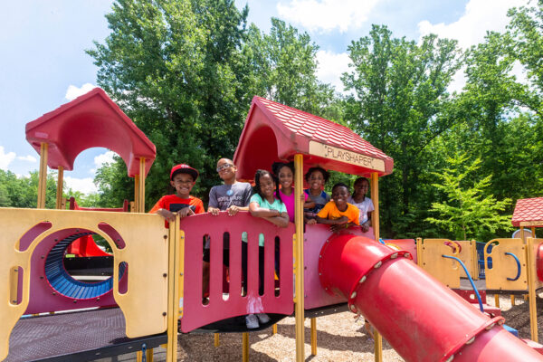 Seven kids pose on the bridge of a red playground structure. Several pathways, slides, and tunnels lead to other play areas.