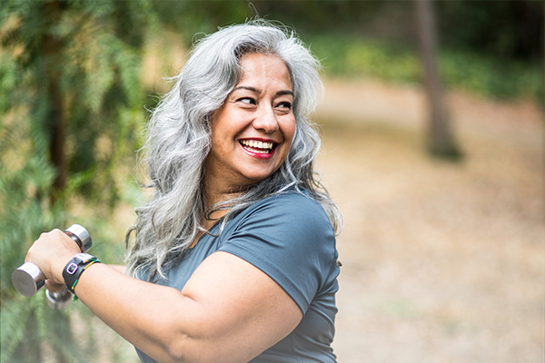 A smiling woman exercises with small silver hand weights outdoors.