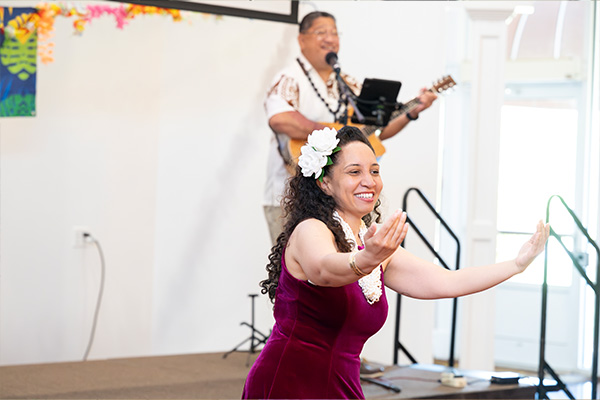 A smiling woman with a large flower in her hair dances in front of a stage on which a man in a Hawaiian shirt is singing and playing guitar.