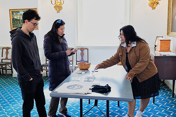 Two visitors speak with a staff member behind a table in a historic room with blue carpet.