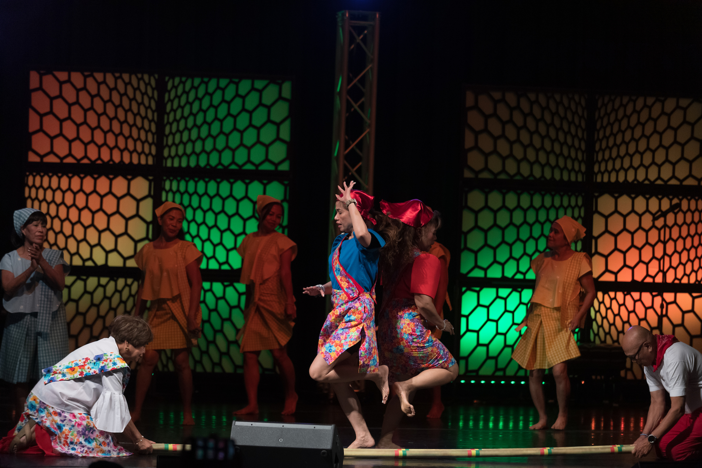 Folk dancers performing a dance on a stage, jumping over a bamboo pole.