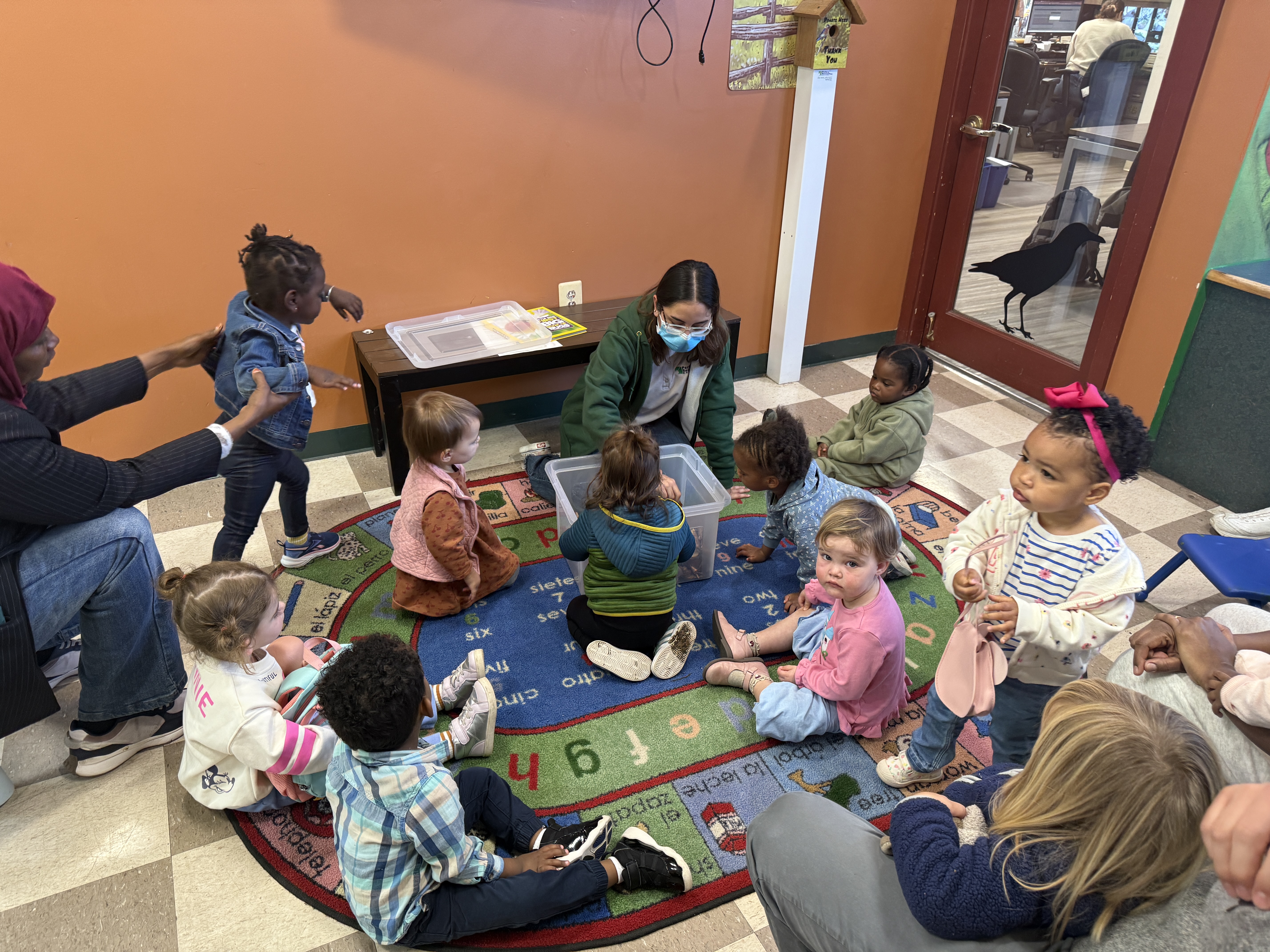 Nine toddlers gather on a colorful rug around a staff member who reaches into a large plastic bin.