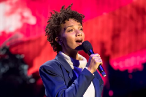 A woman sings into a handheld microphone in front of a red backdrop.