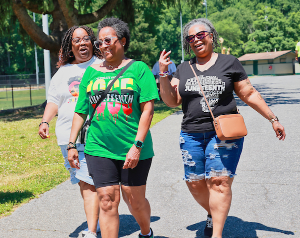 Three people walking along a path on a sunny day. Their shirts all have some graphical variation of the word "Juneteenth"