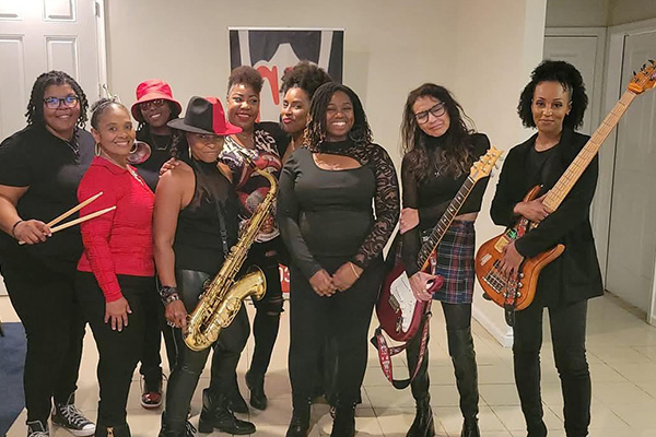 Nine women pose in black and red outfits. Some hold musical instruments, including guitars, a saxophone, and drum sticks.