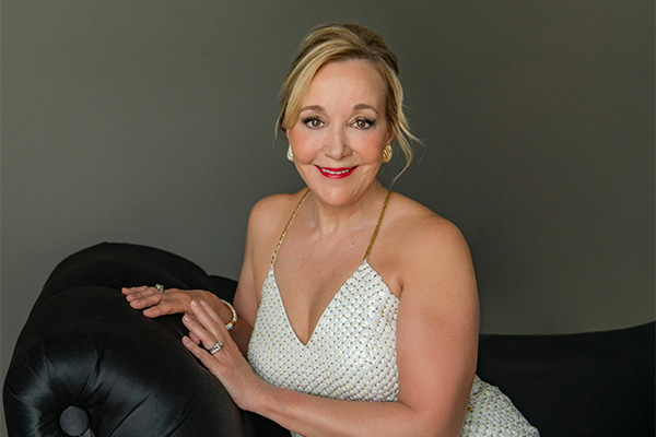 Promotional headshot of a woman in a white evening gown posing on a black settee.