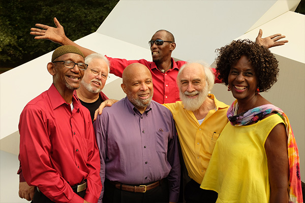 Six smiling adults in solid-color shirts pose in front of a large white sculpture.