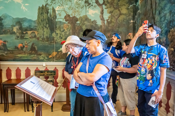 Visitors view a museum exhibit with a large mural, while one person photographs the display.